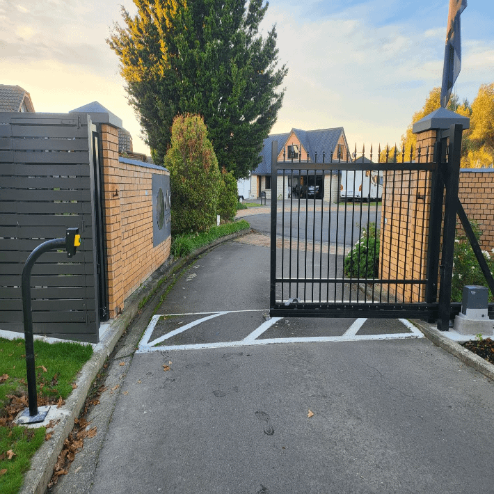 Automatic black metal driveway gate with intercom system opening onto residential street at sunset